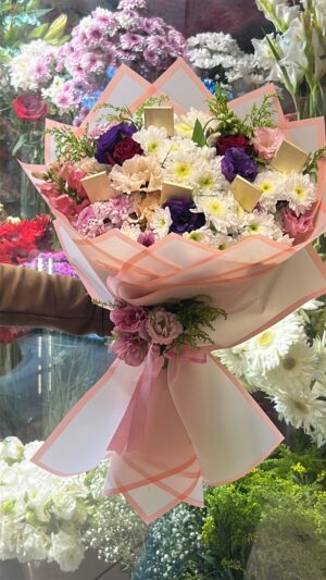 Beyaz, pembe, mor ve krem çiçekler; altın tonlu paketli çikolata detayları, pembe-beyaz ambalaj ve pembe kurdele