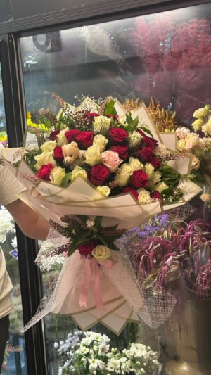 Krem ve kırmızı güller, lilyum tomurcukları ve açık renk çiçekler; pembe tonlu ambalaj