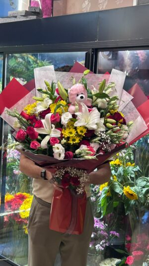 Kırmızı güller, beyaz çiçekler ve sarı dokunuşların koyu kırmızı ambalajla buluştuğu, pembe peluş detaylı buket