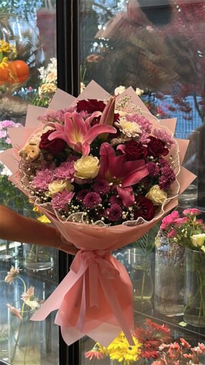Pembe lilyumlar, krem güller ve pembe-mor çiçekler; pembe ambalaj, küçük peluş ve dekoratif ince detaylar