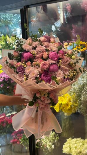 Pembe güller, mor küçük çiçekler ve açık tonlu dokunuşlar; pembe ambalajda yoğun çiçek sunumu