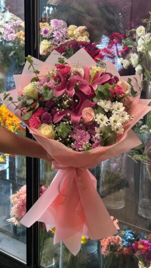 Pembe lilyumlar, pembe ve krem güller, beyaz ve mor çiçekler; geniş pembe ambalaj ve pembe kurdele