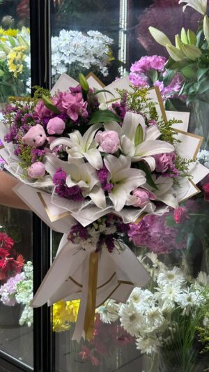 Beyaz ve pembe lilyumlar, açık renkli güller ve mor tonlar; açık ambalajlı karma buket