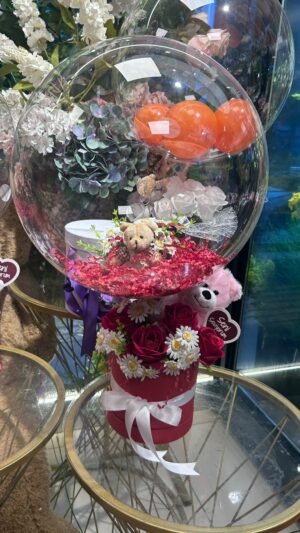 Kırmızı ve beyaz çiçeklerin yer aldığı kutu; üst bölümde şeffaf balon, kalp biçimli detaylar ve peluş