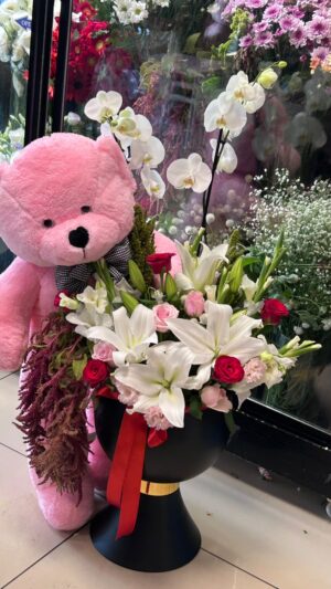 Beyaz ve pembe lilyumlar, kırmızı güller ve beyaz orkide çiçekleri; siyah ayaklı kap ve büyük pembe peluş